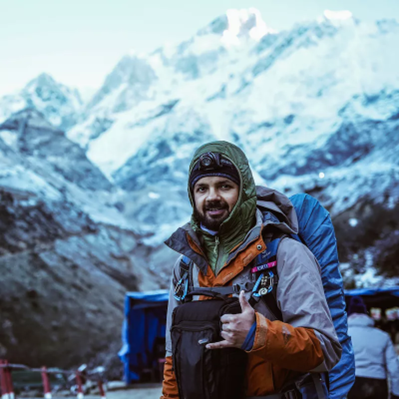 The image shows a man in hiking gear, smiling at the camera with a mountain range in the background. He is wearing a jacket, a headlamp, and a backpack. The mountains are covered in snow, and the overall scene suggests a cold, outdoor environment. The man seems to be enjoying the hike.